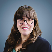 Professional head-and-shoulders portrait of a woman with long brown hair and glasses, smiling in a dark blazer against a blue-gray studio backdrop.