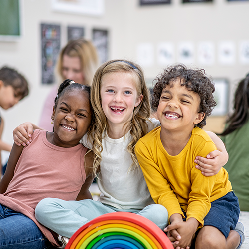 Three young children sitting close together in a classroom, smiling and hugging with a colorful wooden rainbow toy in front of them.
