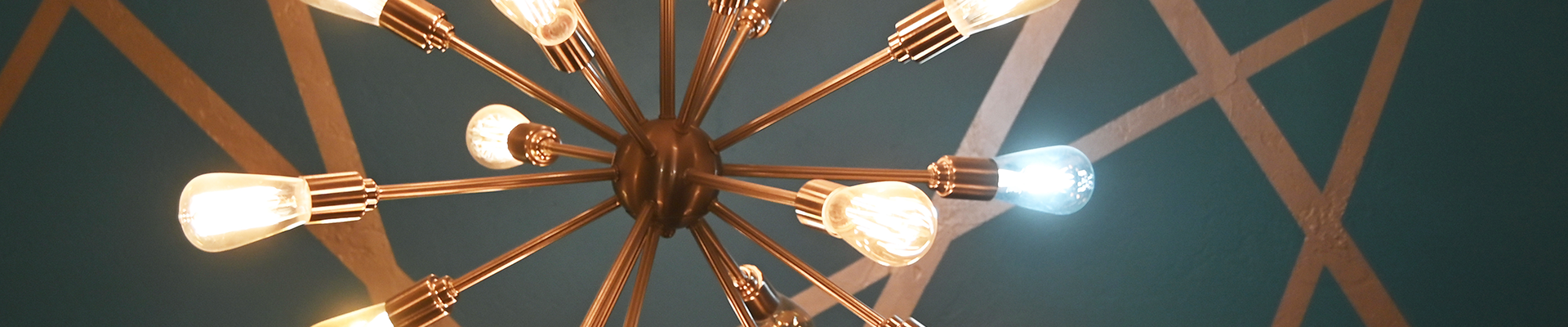 Modern chandelier with multiple exposed bulbs extending from a central hub, viewed from below against a teal ceiling with geometric gold lines.