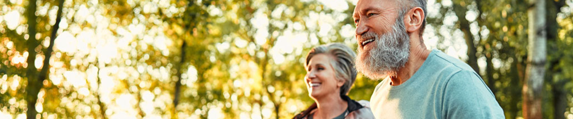 Happy older couple walking through a sunlit forest trail, smiling and enjoying a nature stroll.