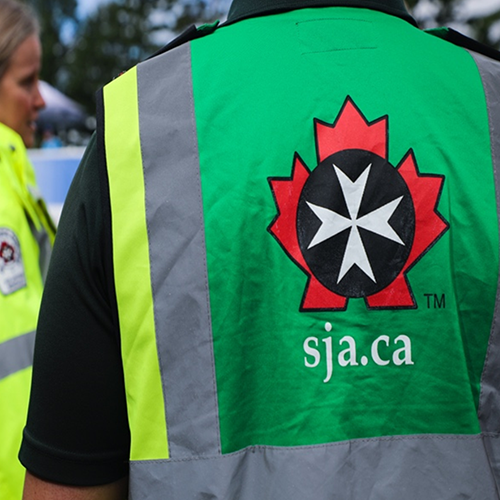Person wearing a high-visibility vest with the St. John Ambulance Canada logo and website (sja.ca) printed on the back, speaking to another individual in uniform.