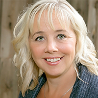 Portrait of a smiling woman with light blonde hair in loose waves, wearing a blue knit sweater, in front of a rustic wood background.