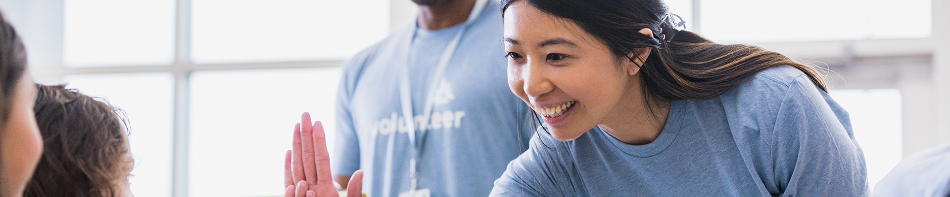 Smiling volunteer in a light blue T-shirt leans in to give a high five to a child during a community service event.