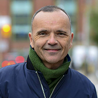 Outdoor portrait of a smiling man in a blue jacket and green scarf, standing on a city street with a blurred background.