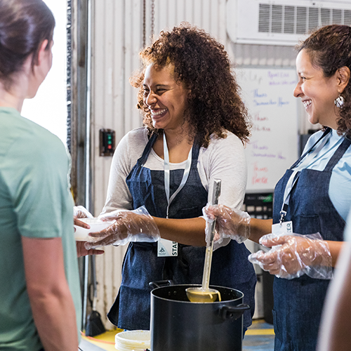 Two cheerful volunteers wearing aprons and gloves serve food from a large pot to a woman at a community meal program.