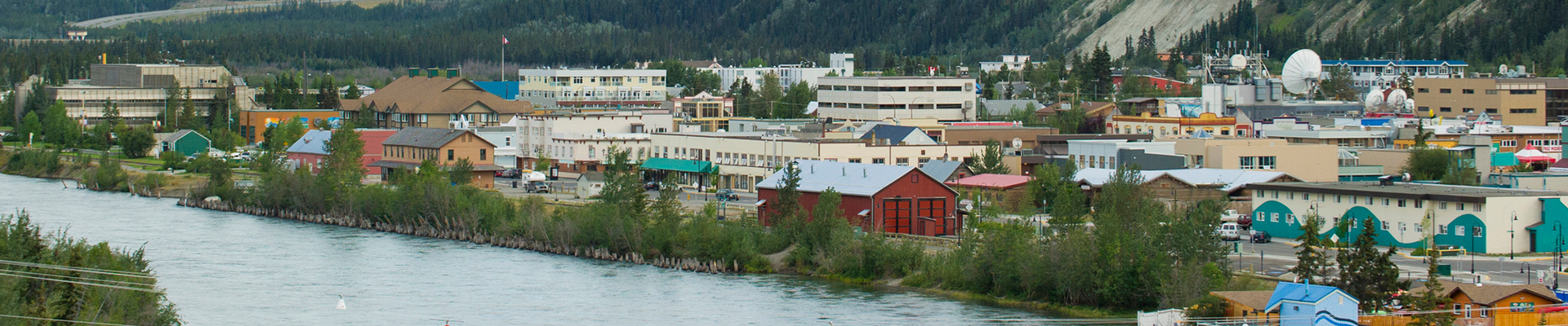 Panoramic view of downtown Whitehorse, Yukon, with buildings lining the riverbank and forested hills in the background.
