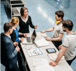 A team of people have a discussion around a worktable with laptops, tablets, and documents