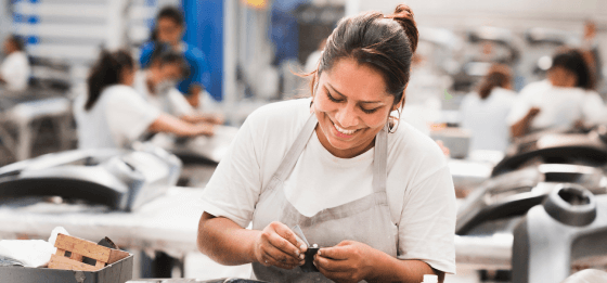 Smiling woman working in a manufacturing plant with other workers visible behind her