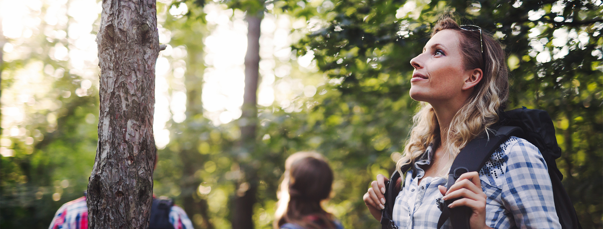 A young woman on a hike in a forest holds her backpack straps and looks up with a smile