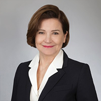 Professional headshot of a woman with short brown hair, wearing a dark blazer and white blouse, smiling against a neutral gray background.