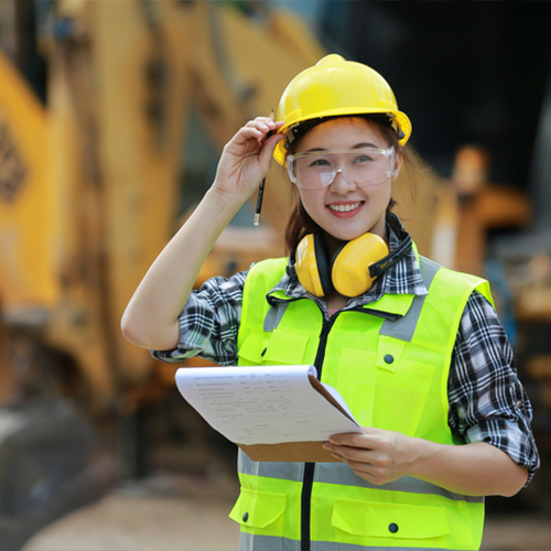Construction-site supervisor in a bright yellow safety vest and hard hat adjusts her helmet and smiles while holding a clipboard, with excavator machinery blurred behind her.