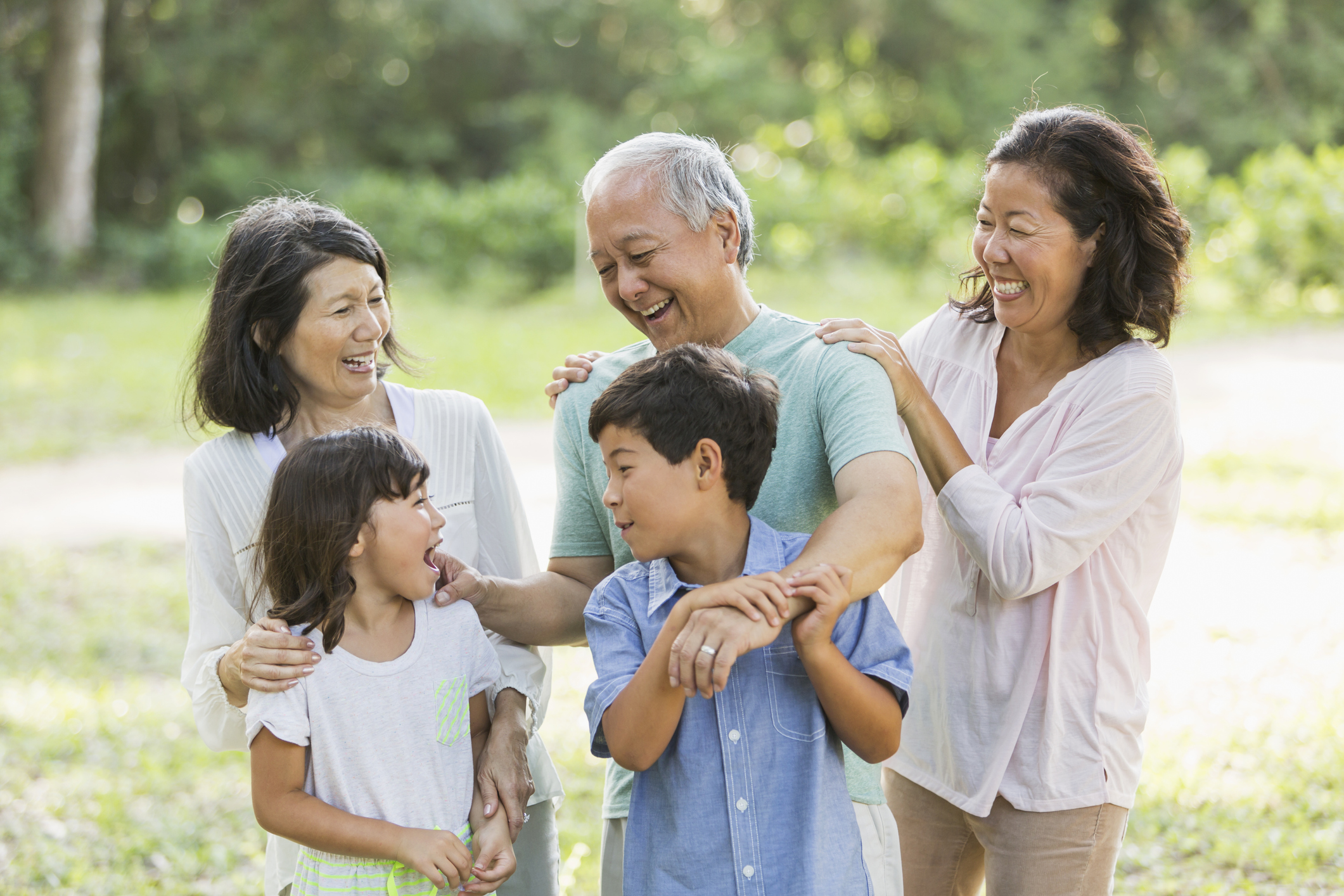 A group of 5 people from a multi-generation Asian family, standinging together in the park.  The mother, in her 40s, and children, ages 6 and 8, are mixed race, Japanese and Caucasian.  The grandparents, standing behind the boy and girl, are Japanese.  They are smiling, laughing and talking.